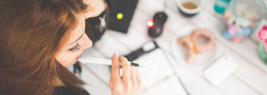 Aerial shot of woman sitting at desk, pen in mouth, looking at planner