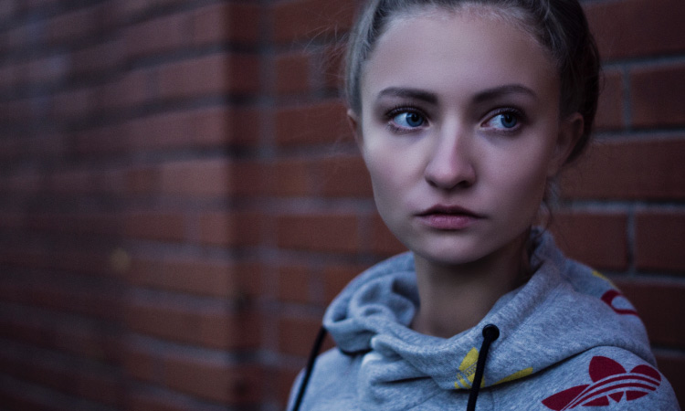 Blonde woman wearing a sweatshirt stands against a red brick wall and looks off anxiously, wondering about cavities