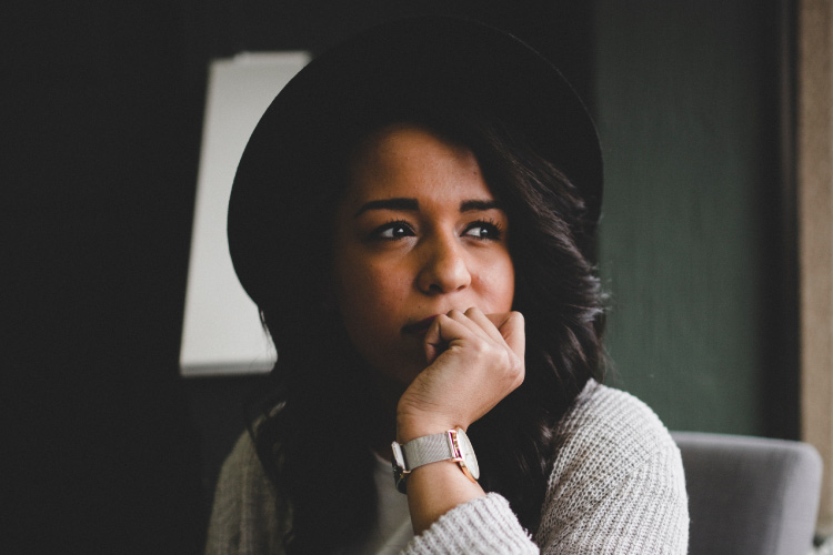 Dark-haired woman wearing a black large-brimmed hat rests her chin on her hand, covering her mouth due to bad breath