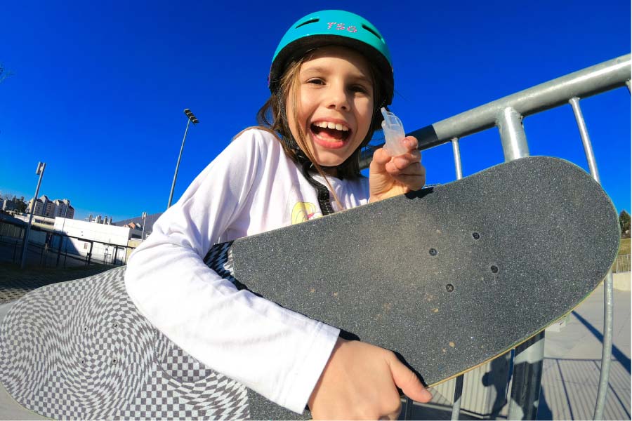 young girl wearing a helmet, skateboard under her arm, holds up her mouthguard and smiles