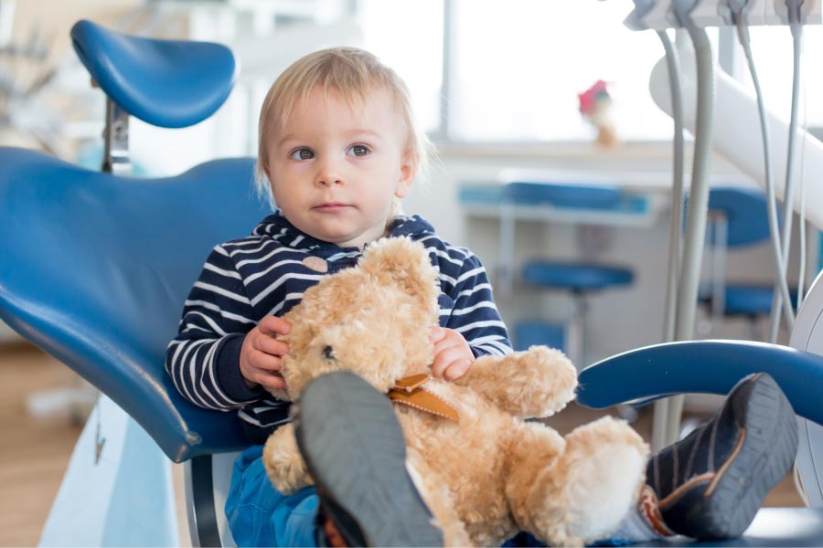 young boy sits in the dentist chair holding a teddy bear